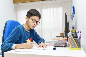 Focused boy preteen doing homework in his room using a laptop and study guides.
