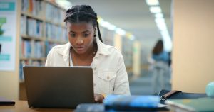 Black woman, student and laptop in library for education, research or studying in school. Girl, computer or reading in university for problem solving, assessment planning or course information on web